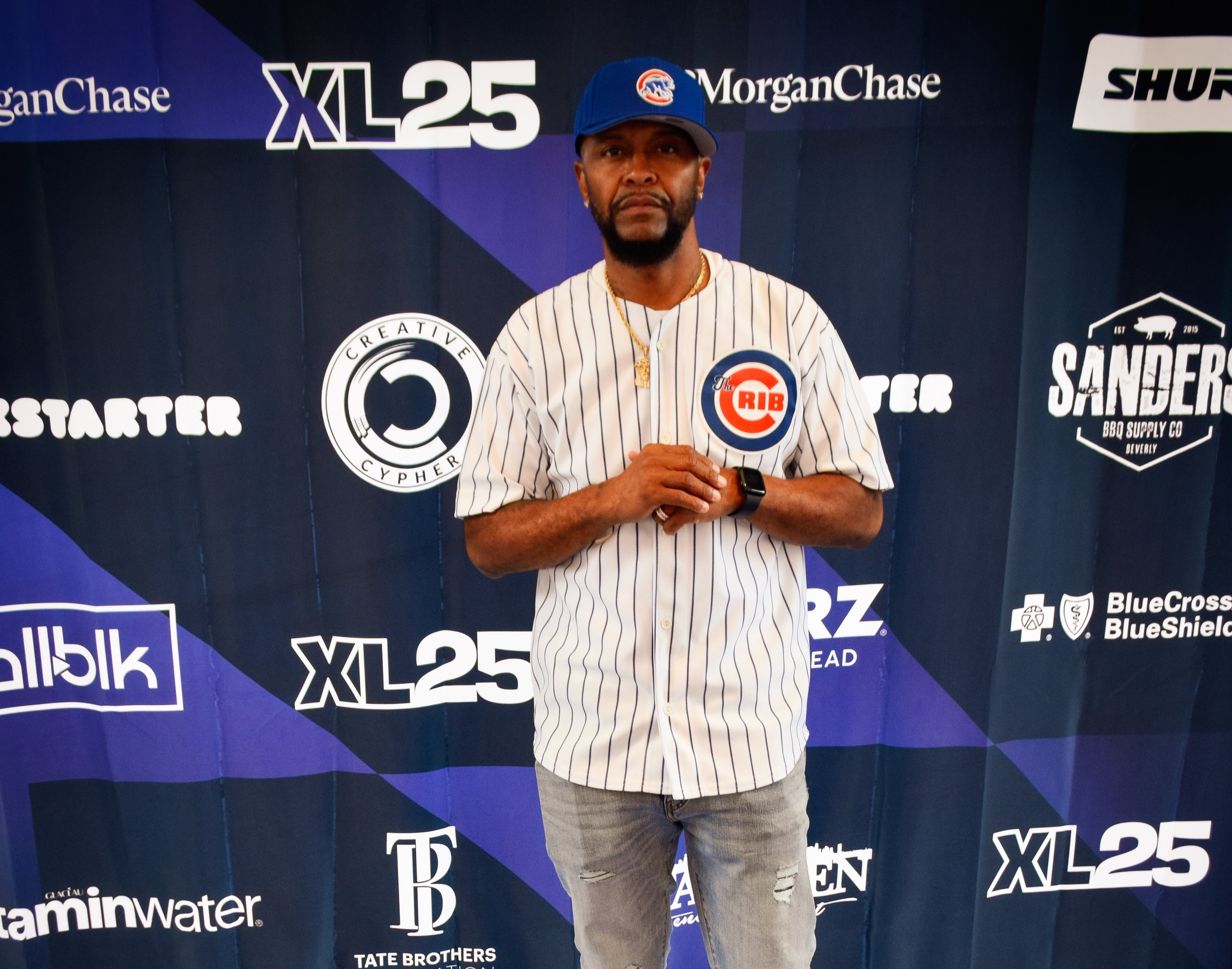 Man wearing a The Crib jersey standing in front of a backdrop with various logos.