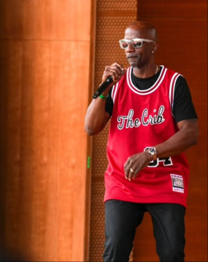 Man wearing a red 'The Crib' jersey holding a microphone against an orange wall.
