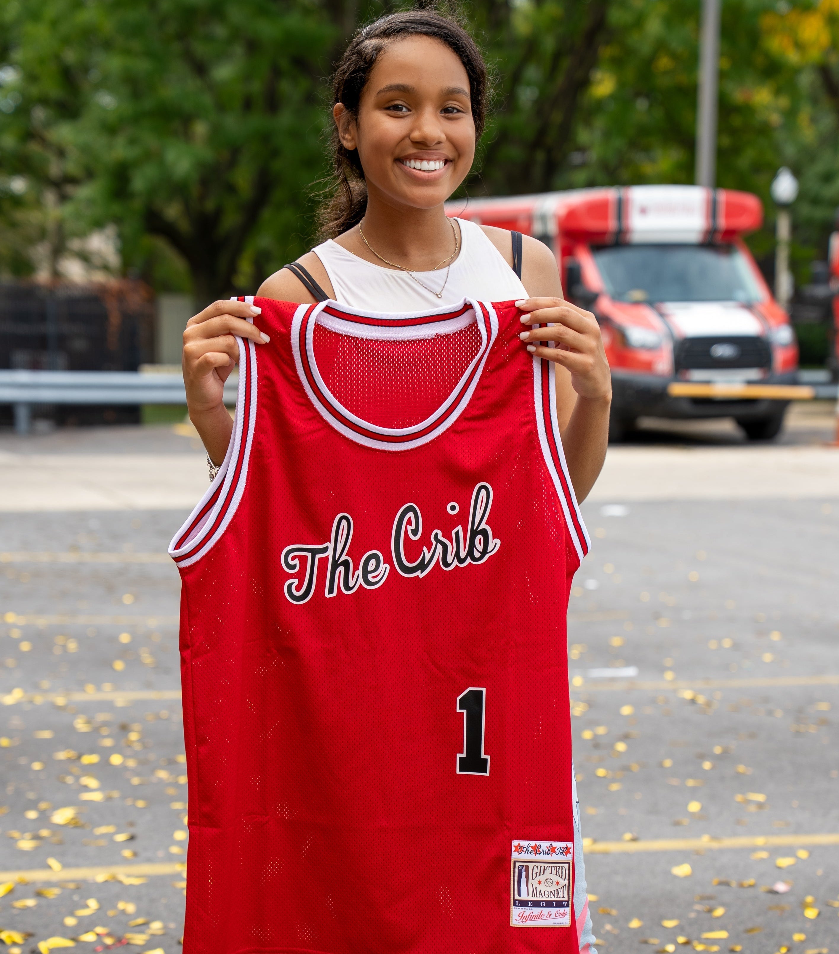 Young lady holding a red basketball jersey with 'The Crib' and number 1 on a street background