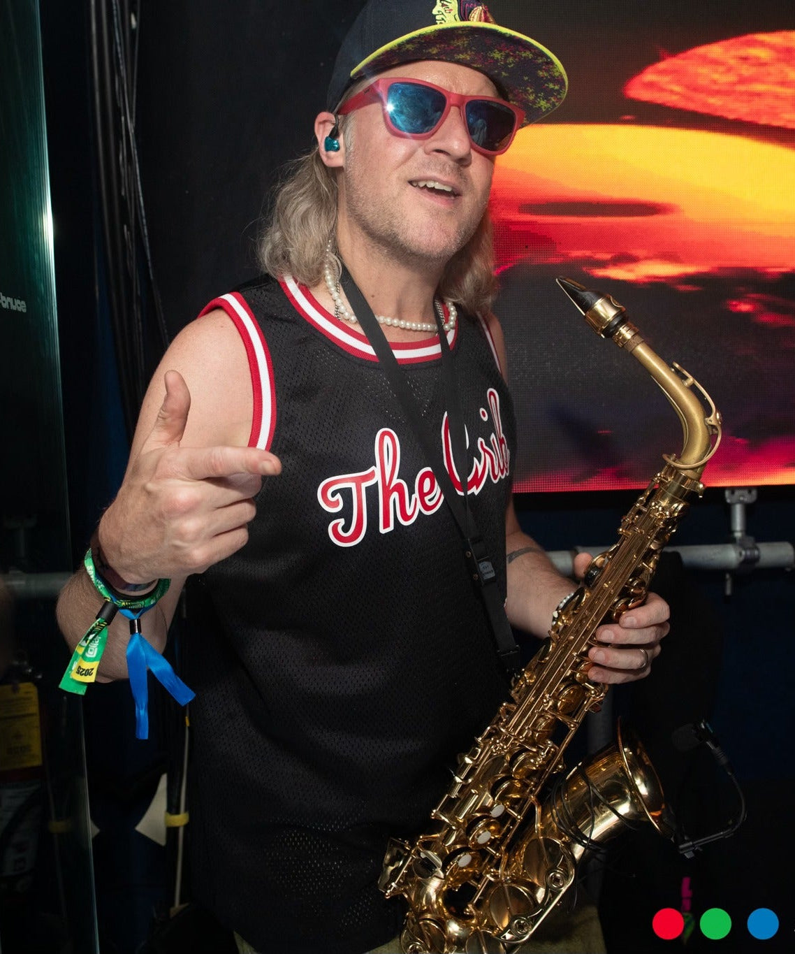 Person holding a saxophone in front of a colorful background with 'The Crib' text on a shirt.