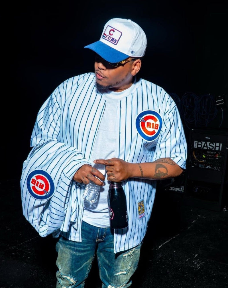 Person wearing a The Crib jersey and cap, holding a bottle, on a dark background